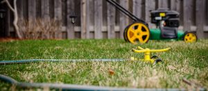 Lawnmower and sprinkler with attached hose sitting on lawn in front of fence.