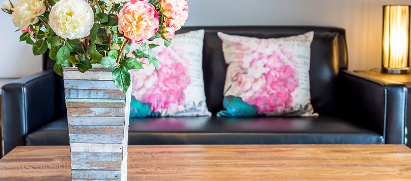 Wooden vase with colorful flowers on table in front of black couch with floral pillows.