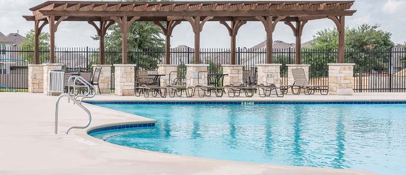 Bright blue community pool with pool chairs and wooden pavilion in background.