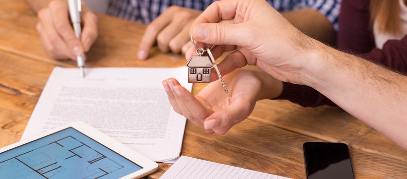 Close up of person handing woman a key with a house while person in background signs closing papers.