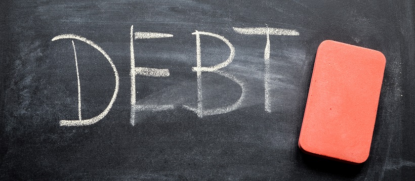 Next to a pink eraser, the word “DEBT” is written on a chalkboard that has been slightly erased.