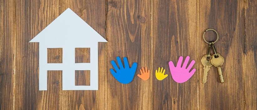 White paper home next to colorful paper hands and keys on a wooden background.