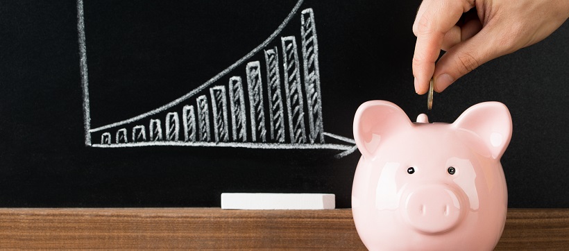 Person’s hand putting coin in piggy bank in front of chalkboard with graph showing upward trend.