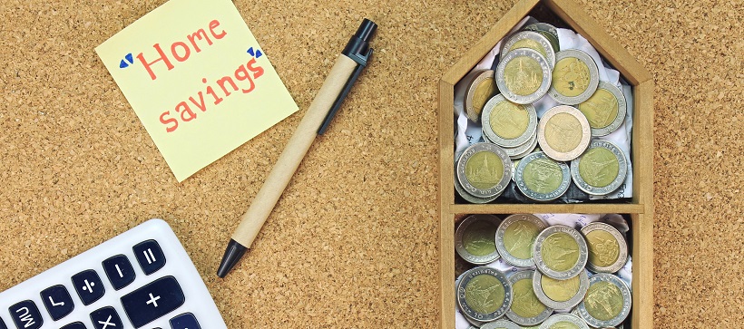 Pen, calculator and sticky note that says “Home savings” next to a wooden model home filled with coins.