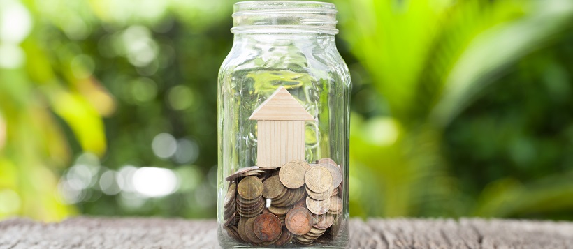 Mason jar sitting on table outside filled with change and a small wooden house.