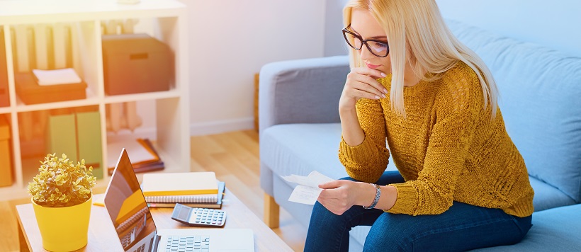 Woman holding receipts on couch in front of laptop and calculator.