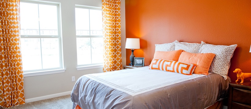 Vibrant bedroom with a gray and white bed, an orange accent wall, orange drapes and orange pillows.