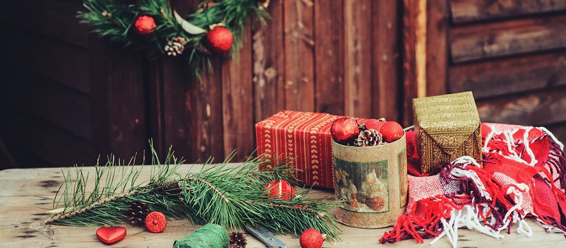 On a wooden table sit Christmas tree branches, a red and white blanket, and gift wrapping materials in assorted holiday colors.