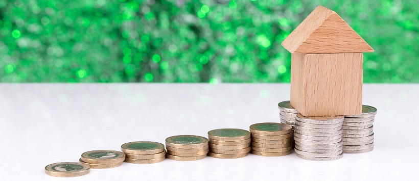 Stacks of coins rising to a pile of coins with a wooden, miniature house sitting on top.