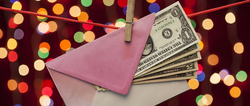  A string and clothespin is holding a pink envelope with dollar bills and Christmas lights in the background.