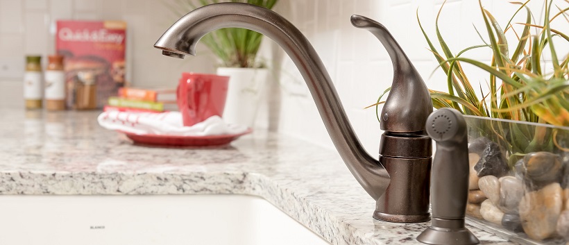 Close-up of a bronze faucet and water sprayer over a sink surrounded by white granite with gray specks.