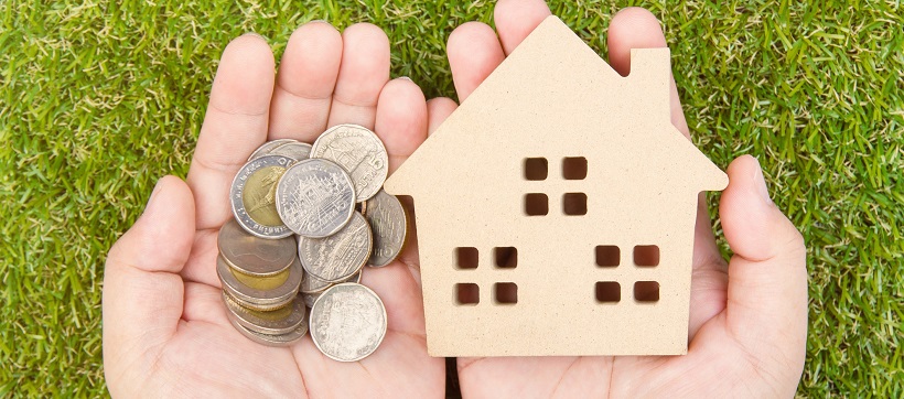 Hands holding coins and a mini wooden home with grass in the background.