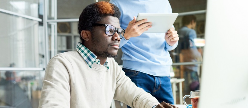 Man sitting at desk working on computer with coworker standing beside him.