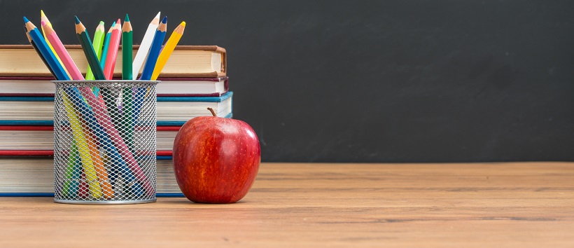 A stack of books, cup of coloring pencils, and an apple sit on a desk with a chalkboard in the background.