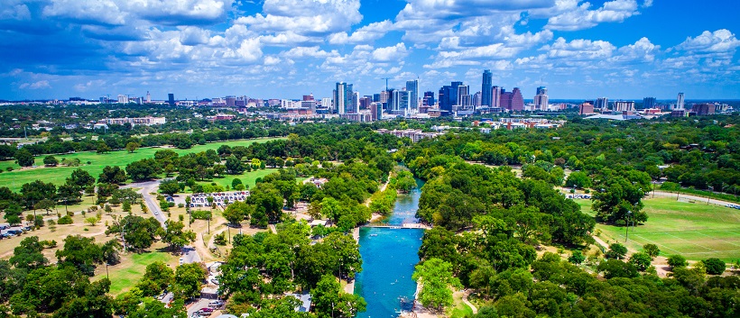 Beneath a blue sky filled with clouds, vibrant trees border a lake traveling towards downtown Austin in the distance.