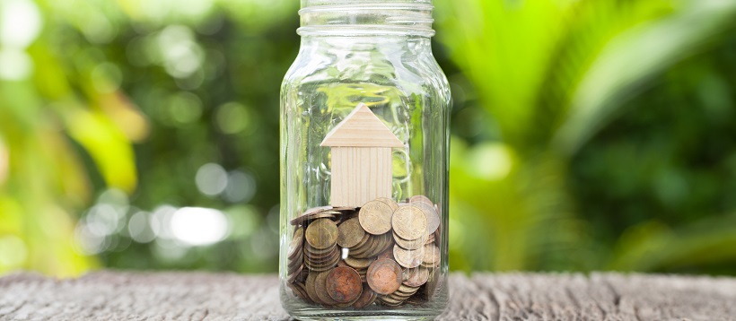 Outside, on a wooden table sits a glass jar with a wooden miniature home on top of coins.