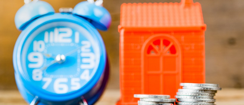 Faded view of a blue clock next to a red model house with silver coins in front.