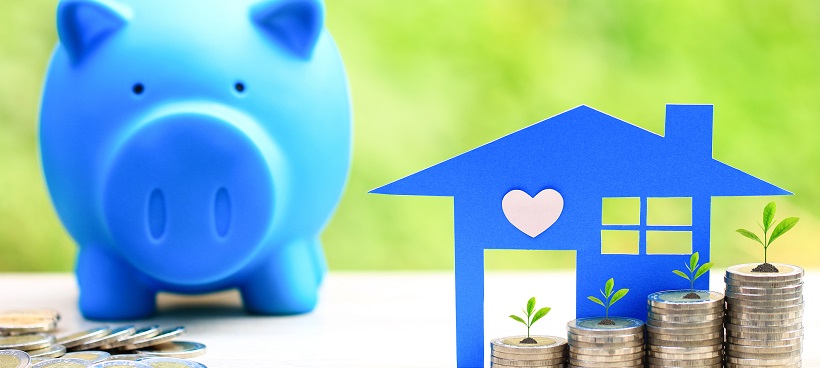 Blue piggybank next to a paper cut-out of a house with stacks of coins in front.