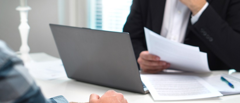 Lender sitting in front of laptop and holding documents from a homebuyer sitting across from him.